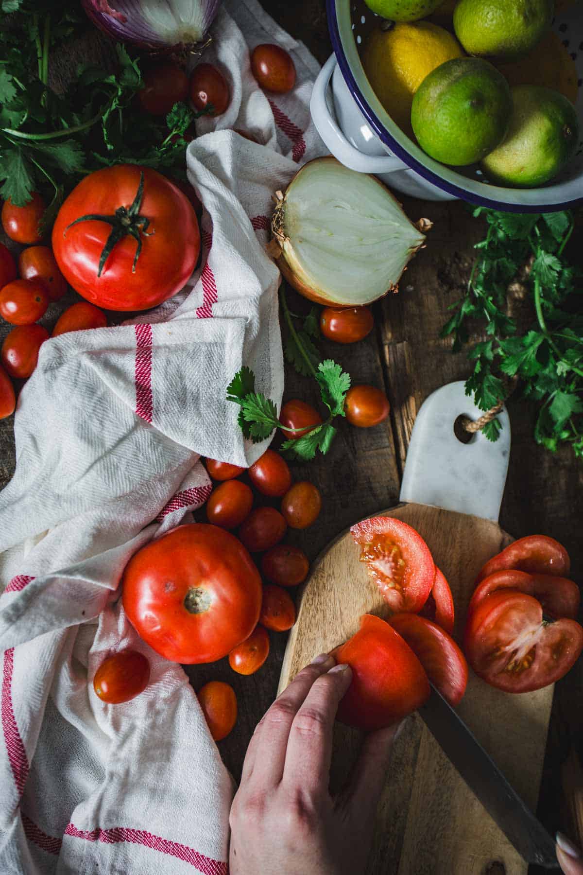 A person is slicing a tomato on a wooden cutting board amidst various whole vegetables and fruits, including lemons, tomatoes, and greens, scattered on a rustic surface with a striped kitchen towel, to prepare