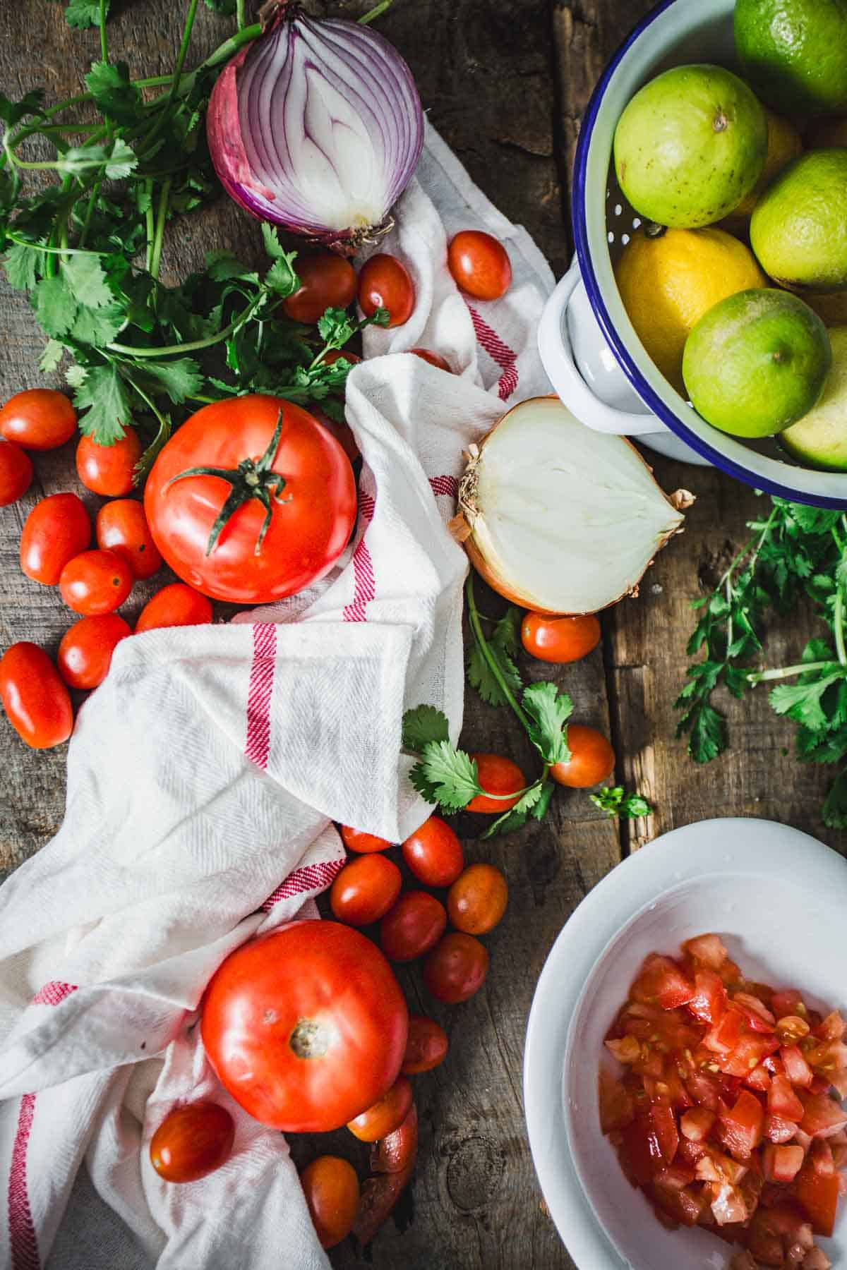 An assortment of fresh vegetables on a wooden surface, including tomatoes, cherry tomatoes, a halved onion, limes, and a sprig of parsley, with a bowl of Colombian empanada sauce