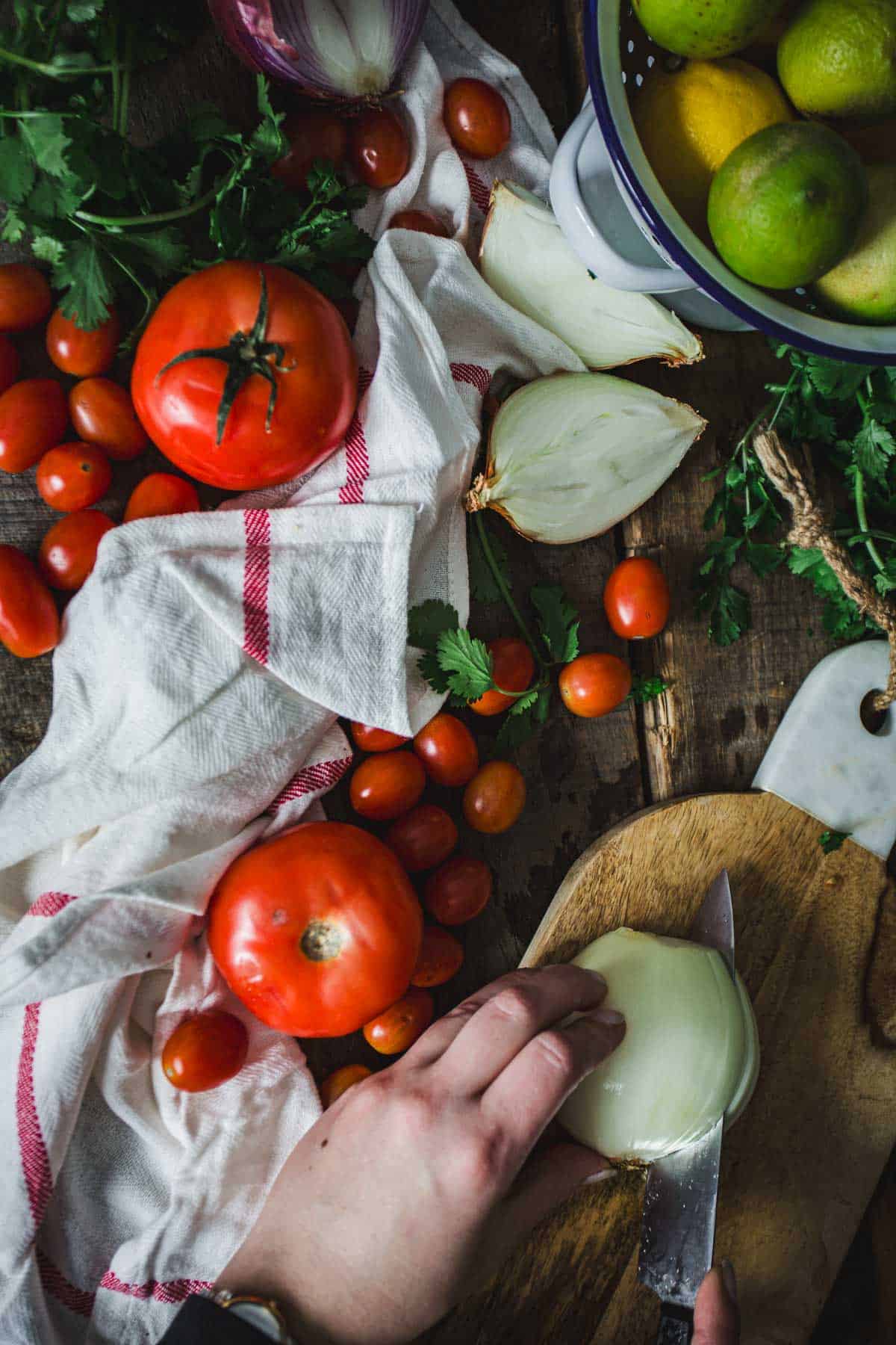 A person slices an onion on a wooden cutting board amidst an array of fresh ingredients including tomatoes, citrus fruits, garlic, and herbs for Colombian empanada sauce.