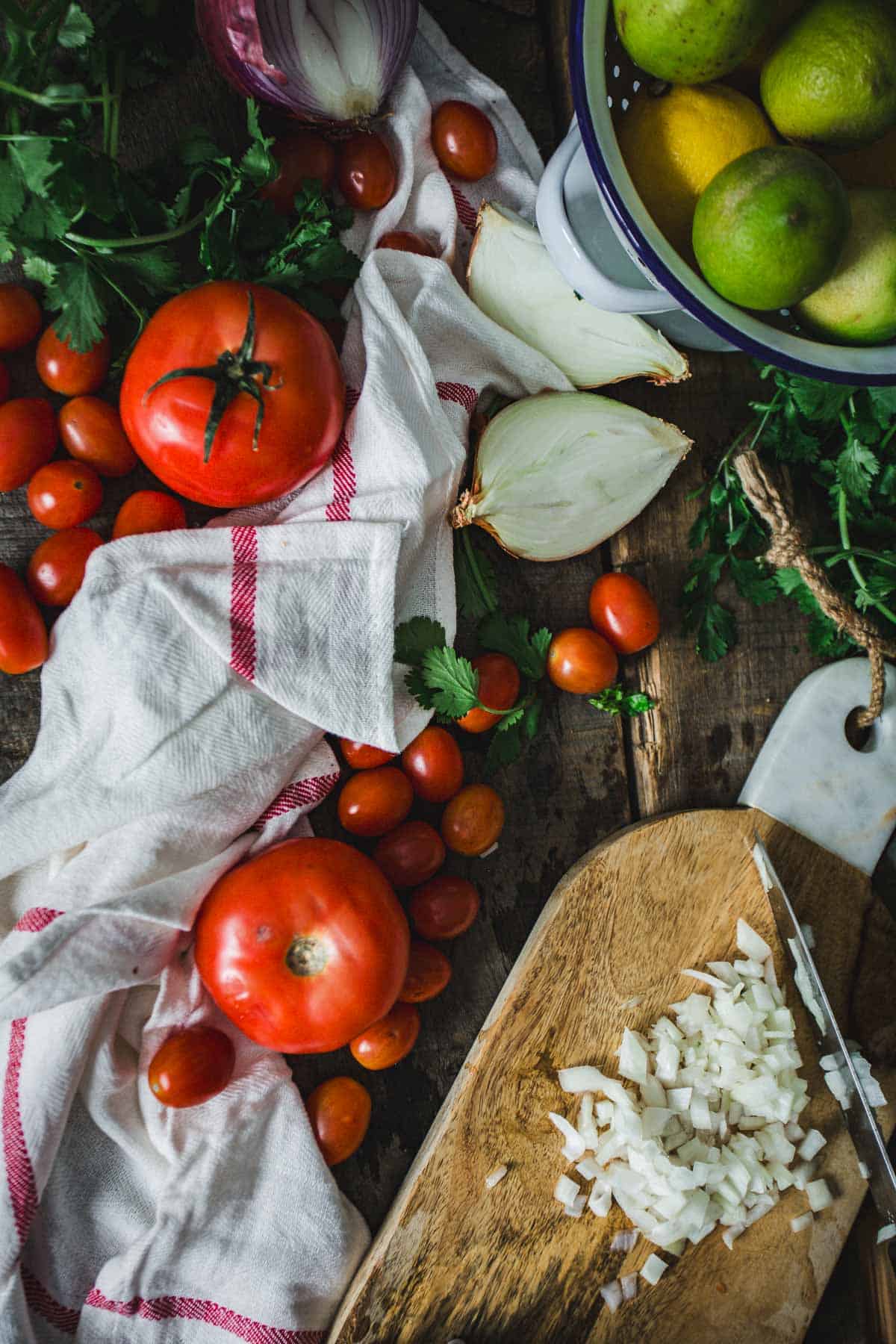 An array of fresh vegetables and citrus fruits on a wooden surface, with chopped onions on a cutting board, Colombian empanada sauce, and a striped cloth nearby.