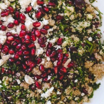 A plate of bulgur salad garnished with pomegranate seeds and crumbled feta cheese. The hearty salad, featuring chopped parsley and finely diced olives, is served on a white plate set upon a wooden surface.