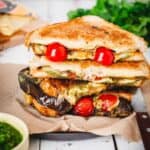 A stacked eggplant grilled cheese sandwich filled with melted cheese, cherry tomatoes, zucchini slices, and eggplant on a piece of brown parchment paper. A bowl of green sauce and leafy greens are partially visible in the background.