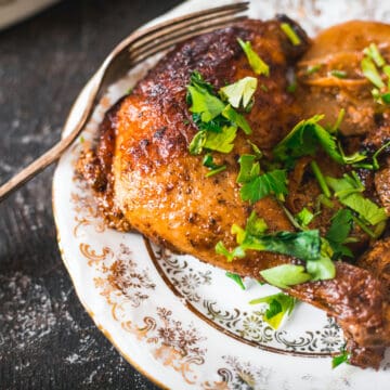 A browned, baked buttermilk chicken leg with crispy skin rests on a decorative plate, garnished with chopped fresh herbs and sliced cooked potatoes. A fork sits at the side, completing the inviting presentation.
