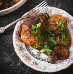 A plate with a seasoned buttermilk chicken leg, sliced potatoes, and fresh parsley garnish, set on a decorative white and gold dish with a fork on a dark textured surface.