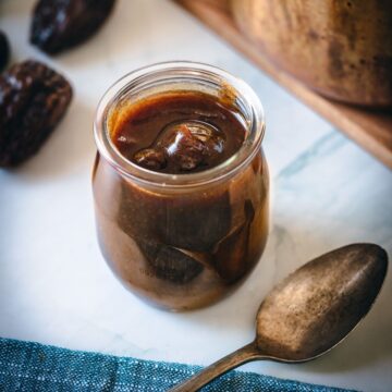 A glass jar of homemade date syrup with a spoon on a kitchen countertop.