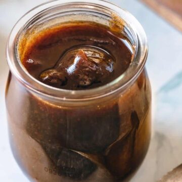 A glass jar filled with dark brown liquid, possibly homemade date syrup with a melting ice cube, resting on a light surface with a blurred background.