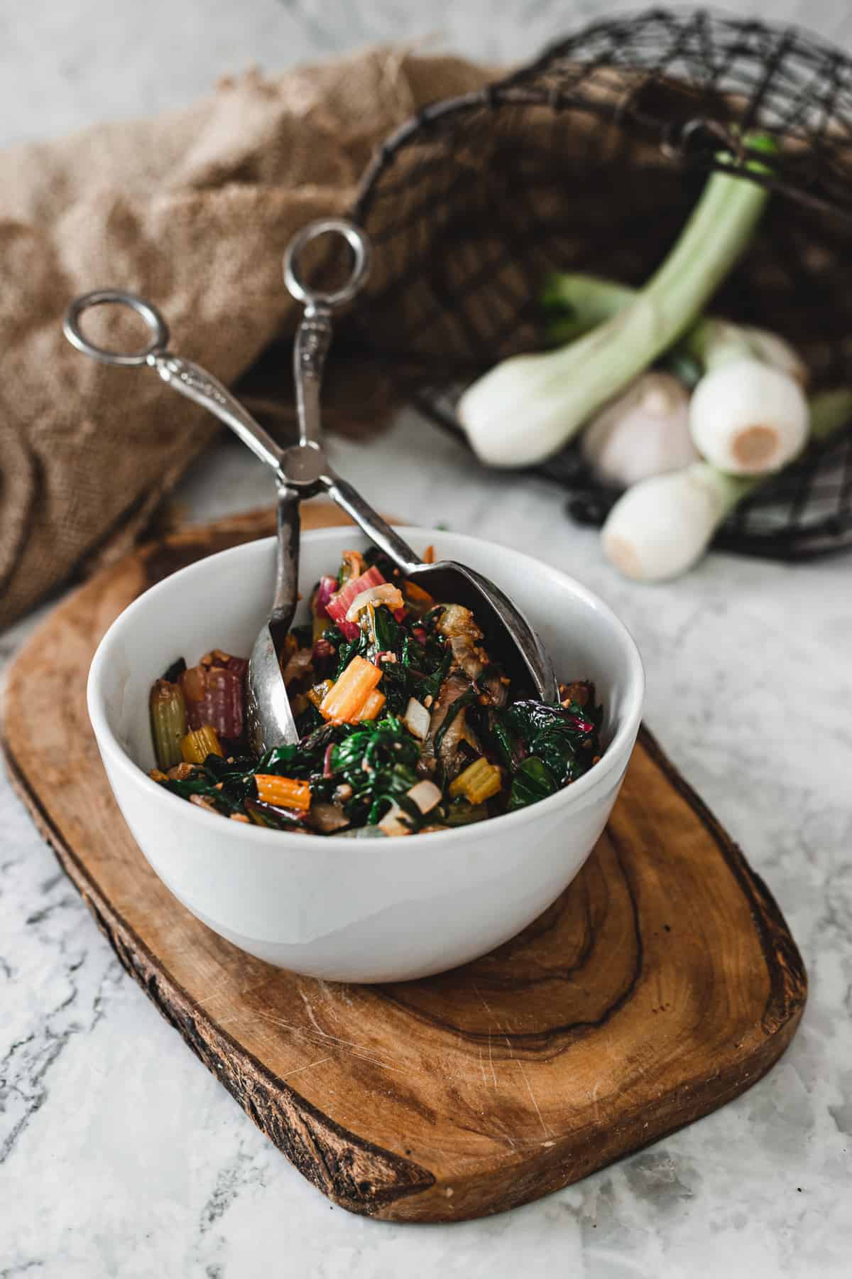 sauteed rainbow chard in bowl with silver thongs on top of small cutting board