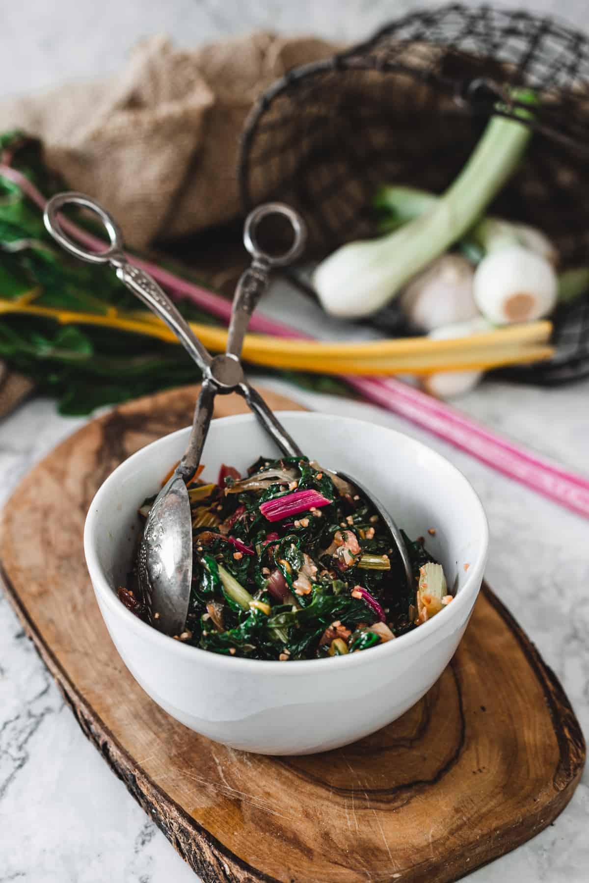 sauteed rainbow chard in bowl with silver thongs on top of small cutting board with onion and rainbow chard in background