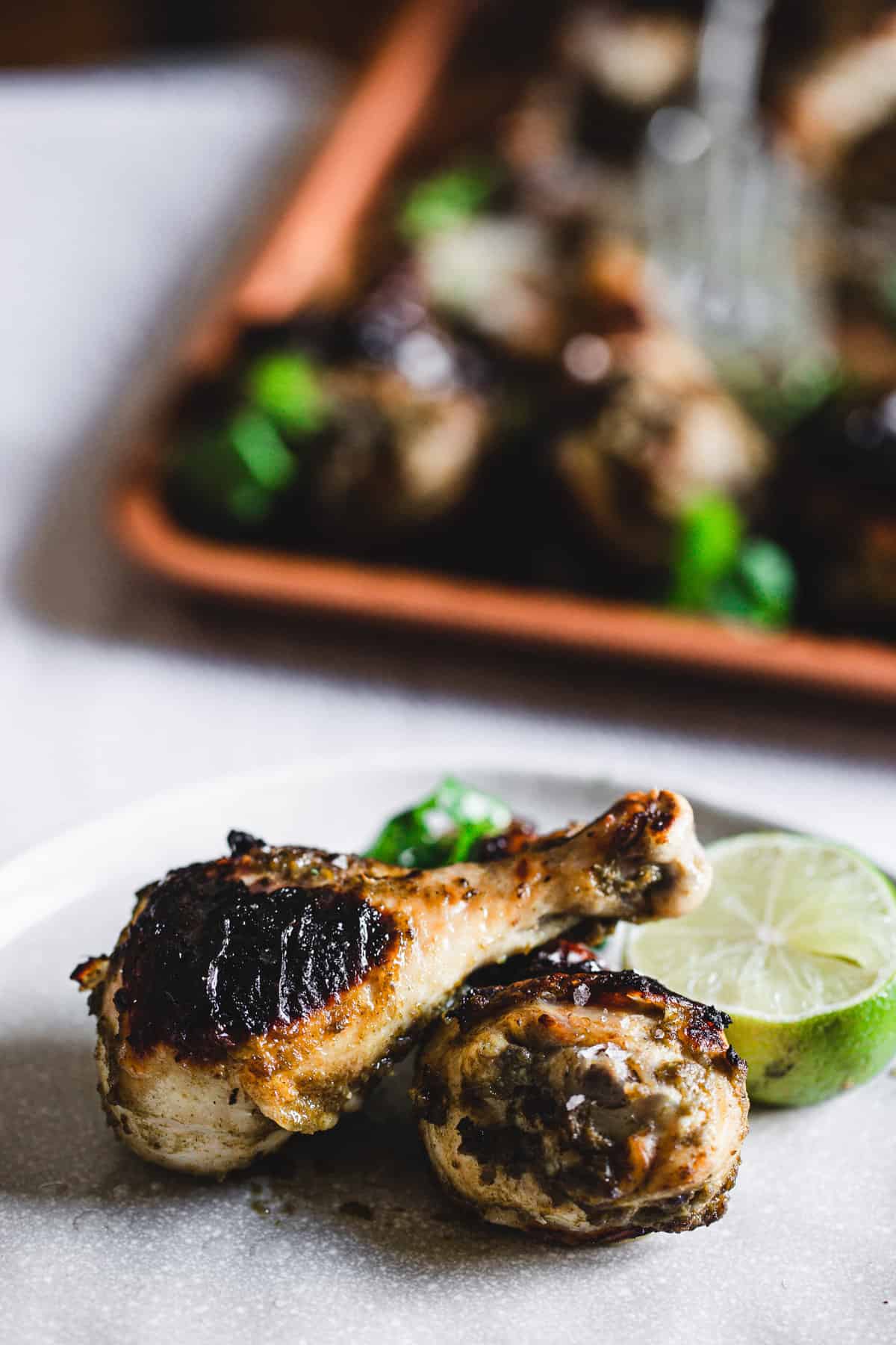 two roasted chicken legs with lime on a plate, tray of chicken drumsticks in background