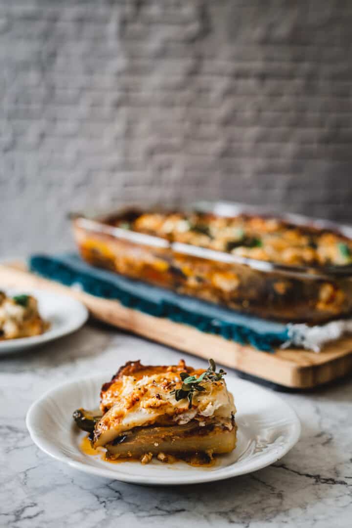 One slice on gluten free vegetarian moussaka on a plate with oregano in the front, and another plate and moussaka pan in the background