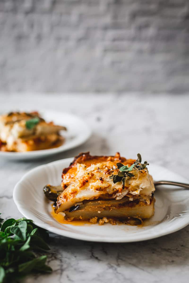 One slice on gluten free vegetarian moussaka on a plate with oregano in the front, and another plate in the background