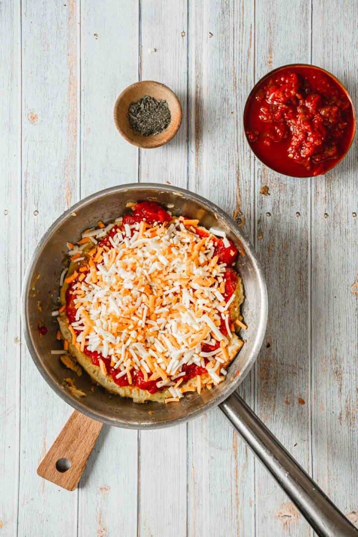 A metal skillet on a wooden surface showcases a matzo brei pizza crowned with shredded cheese and tomato sauce. To the left, a small bowl of herbs complements the dish, while to the right, another bowl brims with chunky tomato sauce.