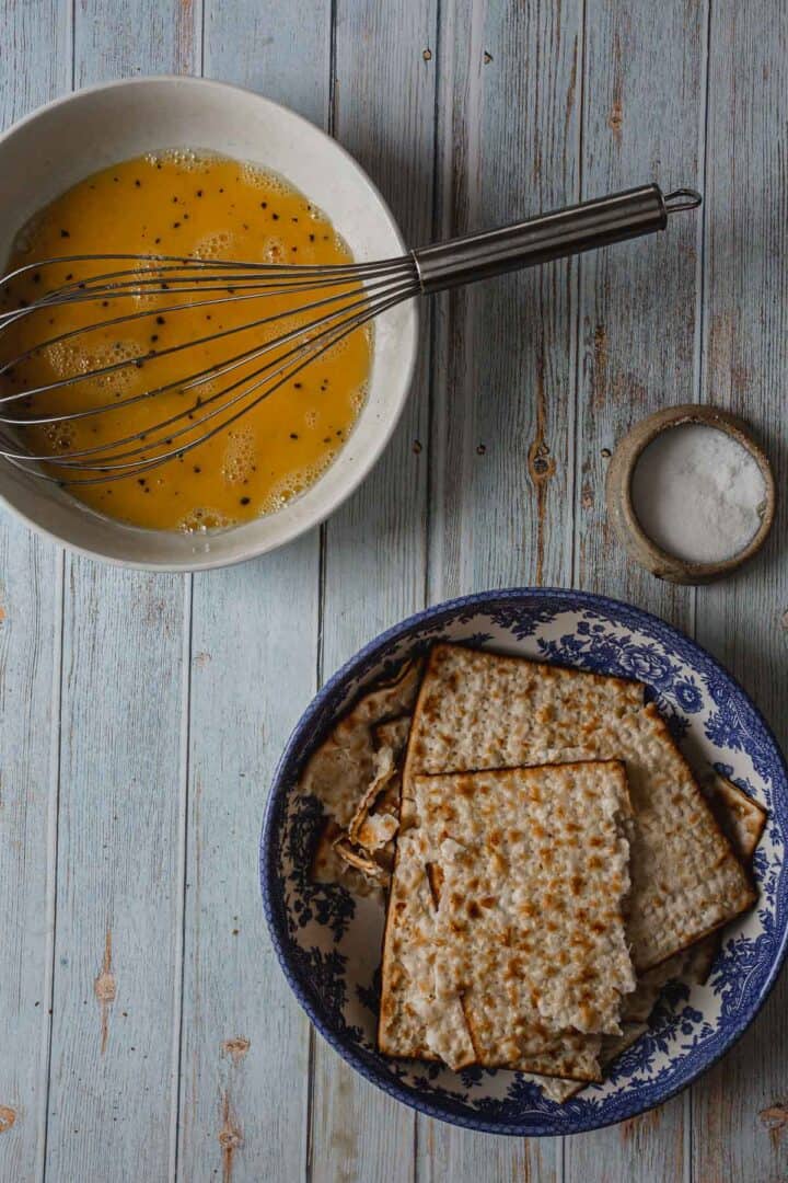 A bowl with a metal whisk and beaten eggs mixed with pepper sits next to a small bowl of salt. Below, a blue patterned dish holds several pieces of matzo bread, like the makings of a savory matzo brei pizza, arranged invitingly on a rustic wooden surface.