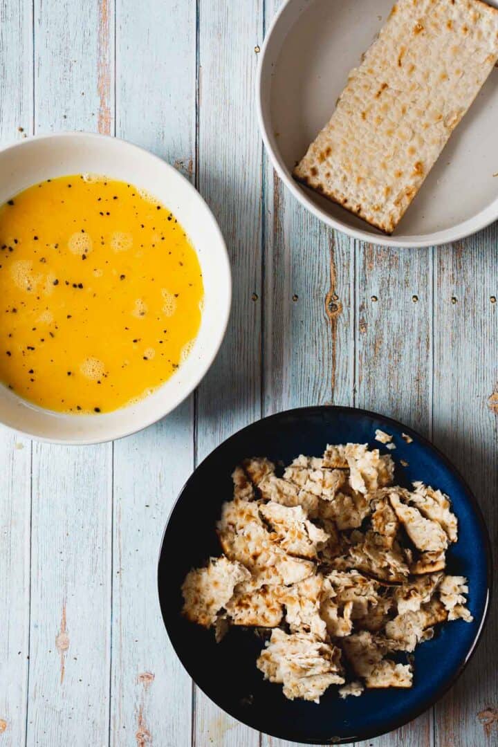 A top-view image of three bowls on a wooden surface. The first bowl contains beaten eggs with black specks, ideal for crafting a matzo brei. The second holds a single rectangular piece of matzah, while the third is filled with crumbled pieces, setting the stage for a creative pizza spin.