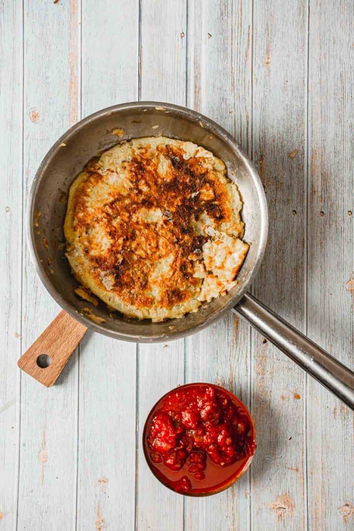 A frying pan with a large, slightly burned pancake reminiscent of matzo brei rests on a wooden surface. Beside it, a small bowl brims with red berry sauce, adding a tangy twist to this breakfast pizza experience.