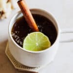 A white mug of aguapanela garnished with a cinnamon stick and lime slice.