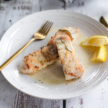 Two portions of white fish in white wine sauce on a white plate with a fork and lemon.