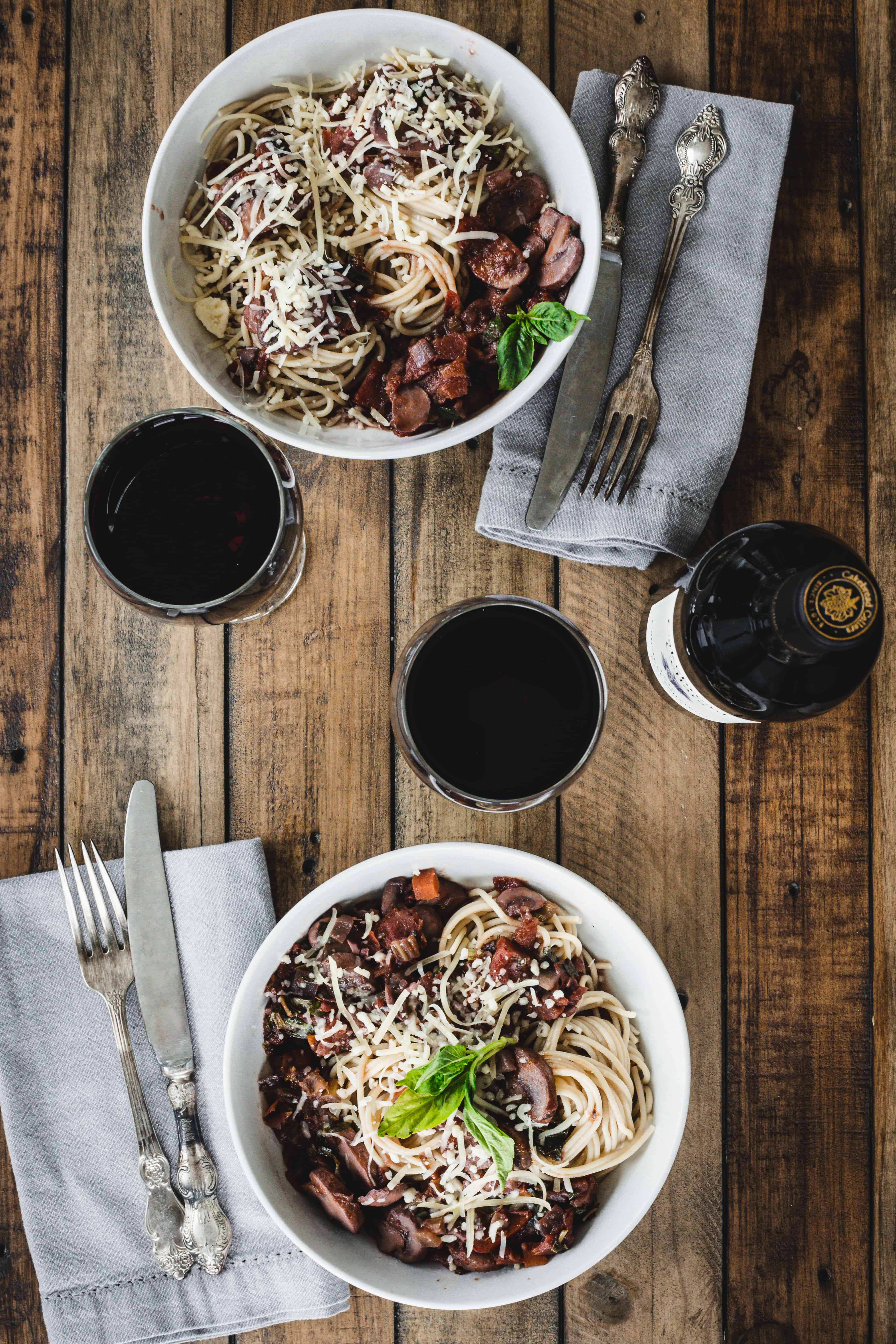 Two bowl of mushroom marinara spaghetti with two glasses of wine and table settings.