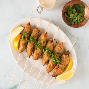 Six chicken kofta kebab skewers on a plate with sauce and cilantro.