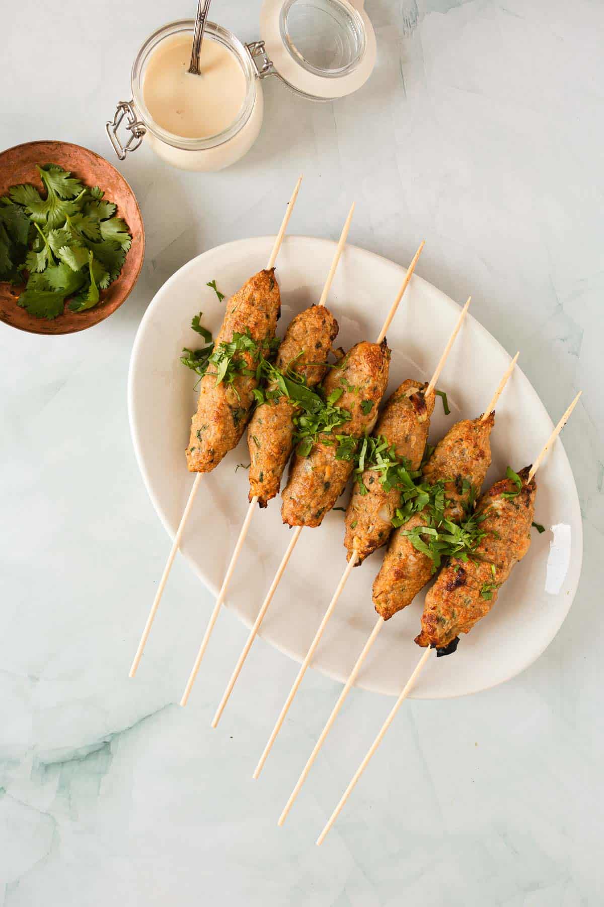 Six chicken kofta kebab skewers on a plate with sauce and cilantro.