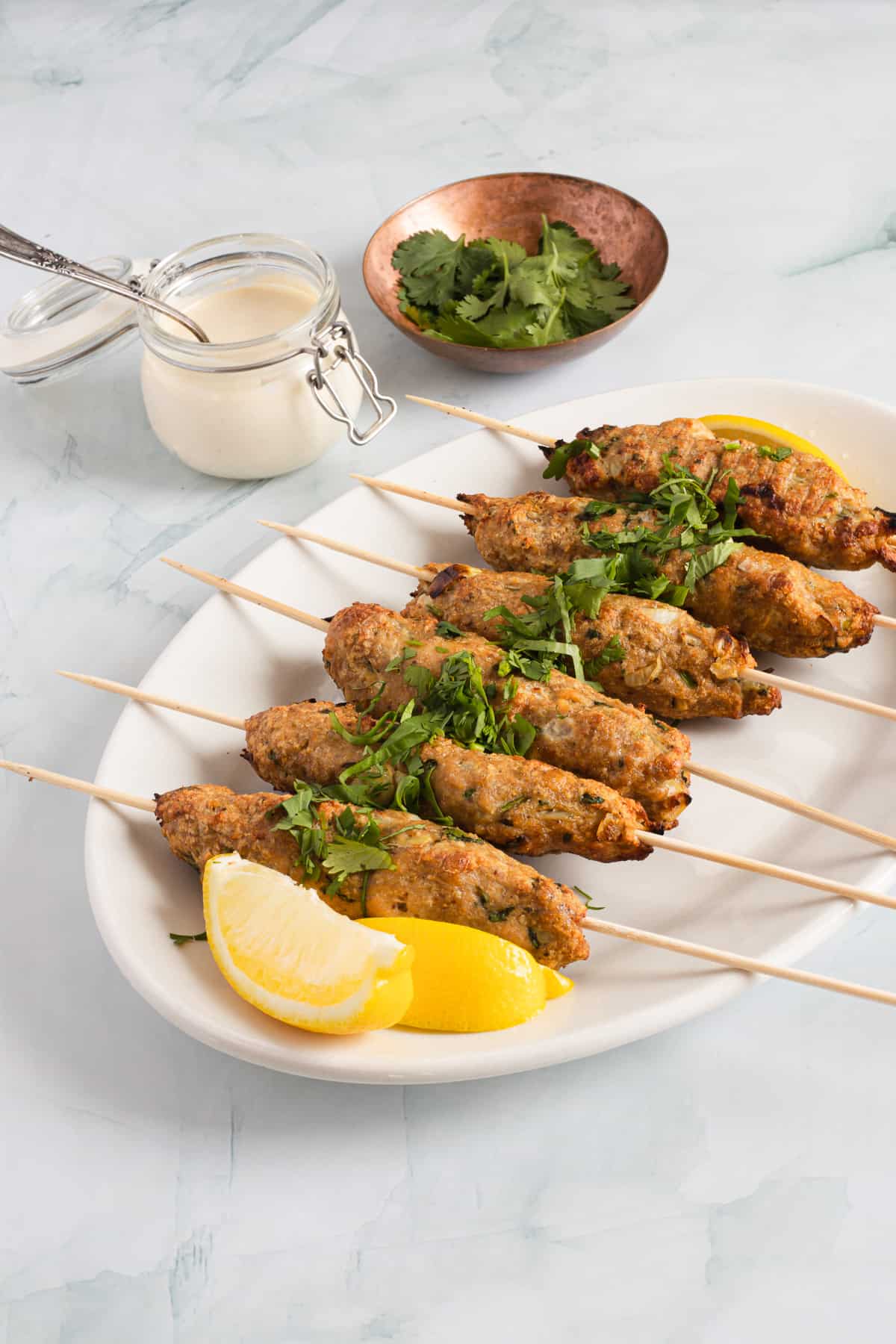 Six chicken kofta kebab skewers on a plate with sauce and cilantro.