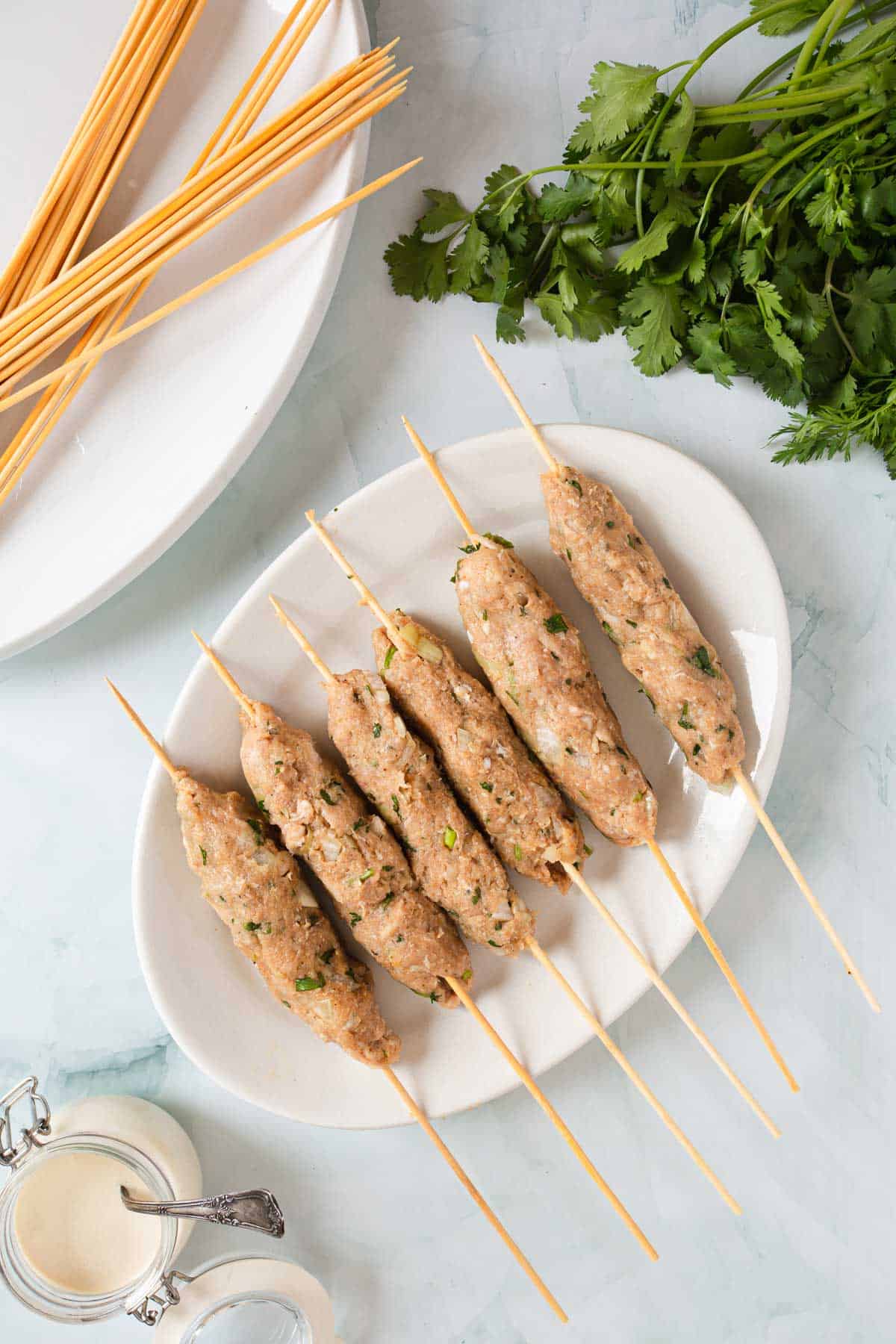 Six raw chicken kofta kebab skewers on a baking sheet with cilantro and skewers.