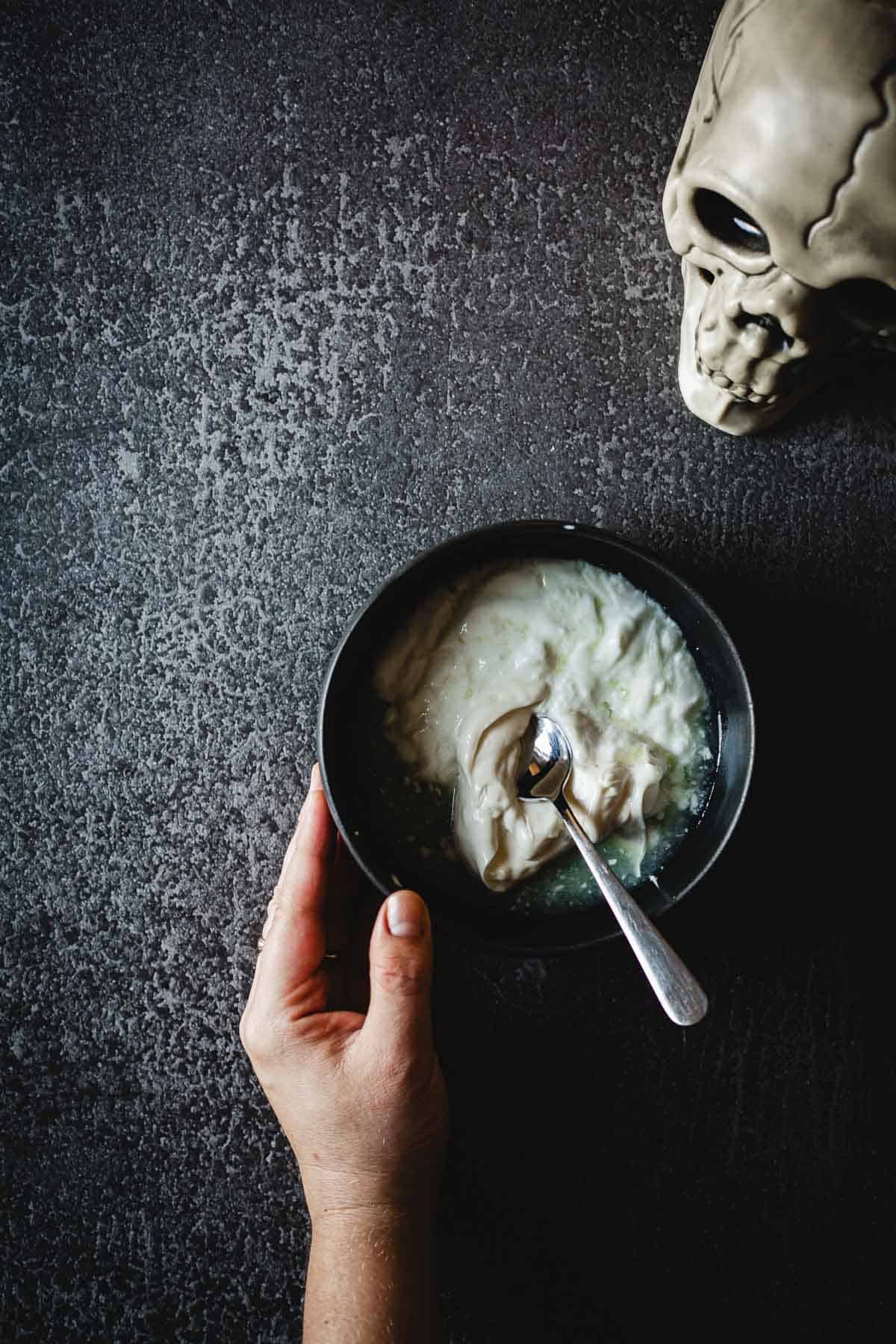 A hand holding a bowl of prepared sauce next to a skull.