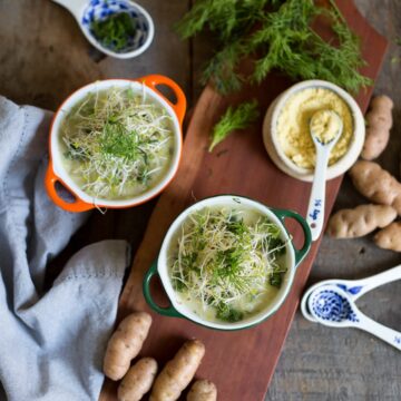 Two bowls of creamy, low sodium potato leek soup garnished with shredded vegetables and fresh herbs sit on a wooden surface. Surrounding the bowls are cloth napkins, baby potatoes, blue and white spoons, and a small dish of powdered seasoning.