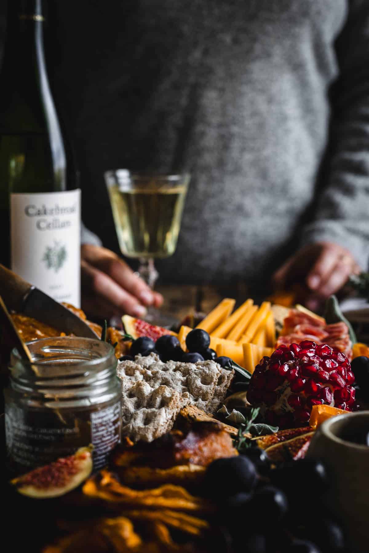 A person holding a bottle of wine next to a platter of cheese and fruit.