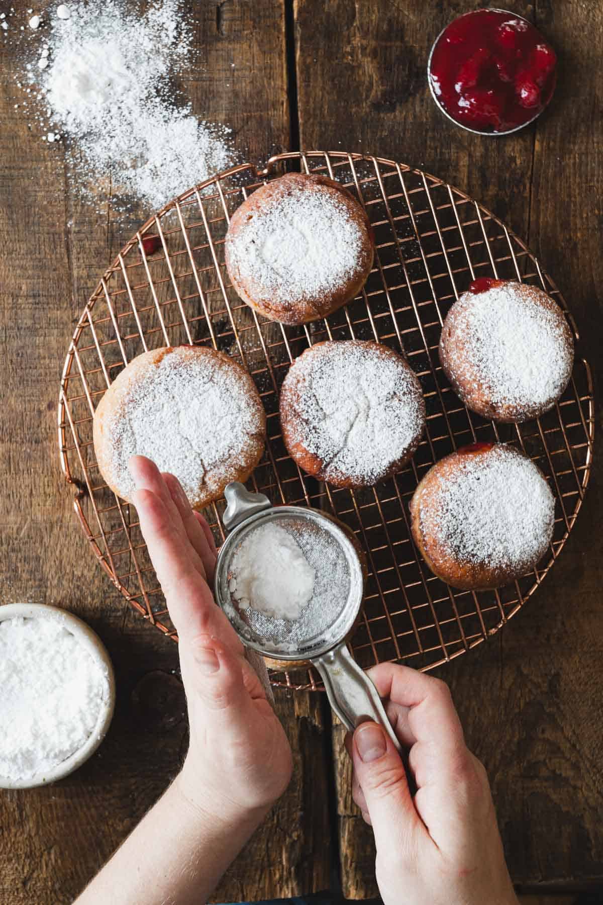 A person is sprinkling powdered sugar on a cooling fried donuts.