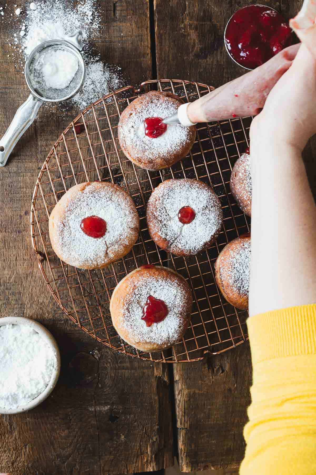 A person is sprinkling powdered sugar on chilled jelly donut on a cooling rack.
