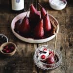 A dish with four wine-poached pears in a red sauce is on a wooden table. In front of it, a glass bowl contains a poached pear half, topped with cream and red berries, with a fork resting on the side. Small bowls with extra ingredients are scattered around.