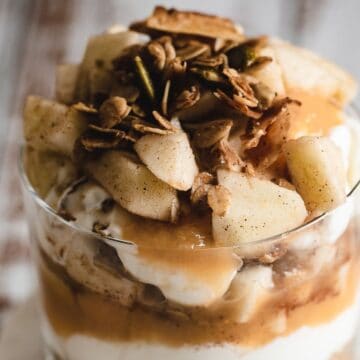 A glass of yogurt topped with apples and pistachios sprinkled with cinnamon.