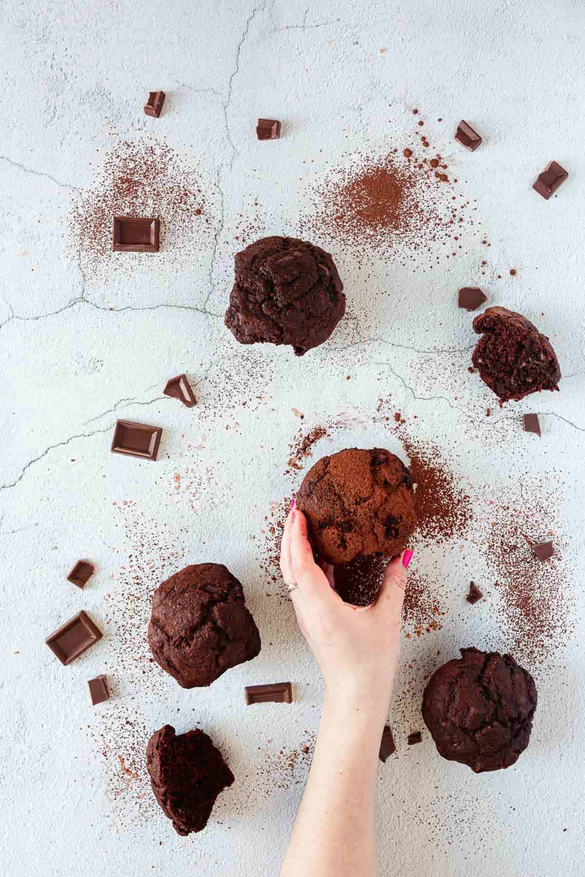 A hand reaching for gluten free triple chocolate muffins on a white surface.