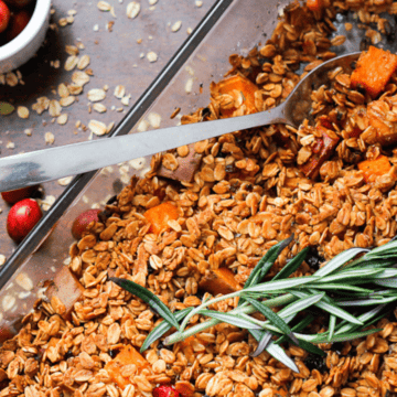 A baking dish filled with oven-baked granola comprising oats, nuts, and dried fruits, garnished with a sprig of rosemary, alongside a small bowl of fresh cranberries. This comfort