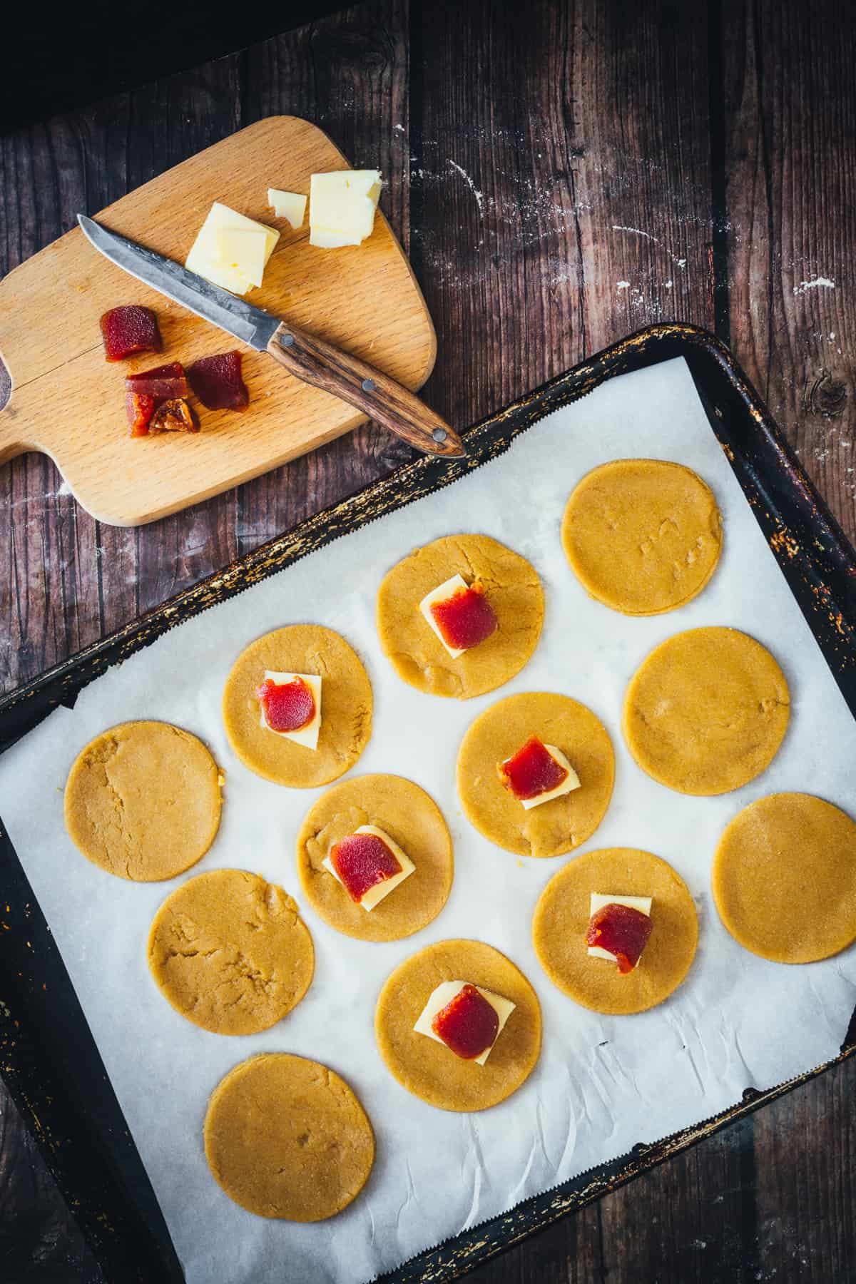 Unbaked cookies with squares of Bocadillo Guava on a parchment-lined baking tray, with a wooden board and chunks of cheese to the side.