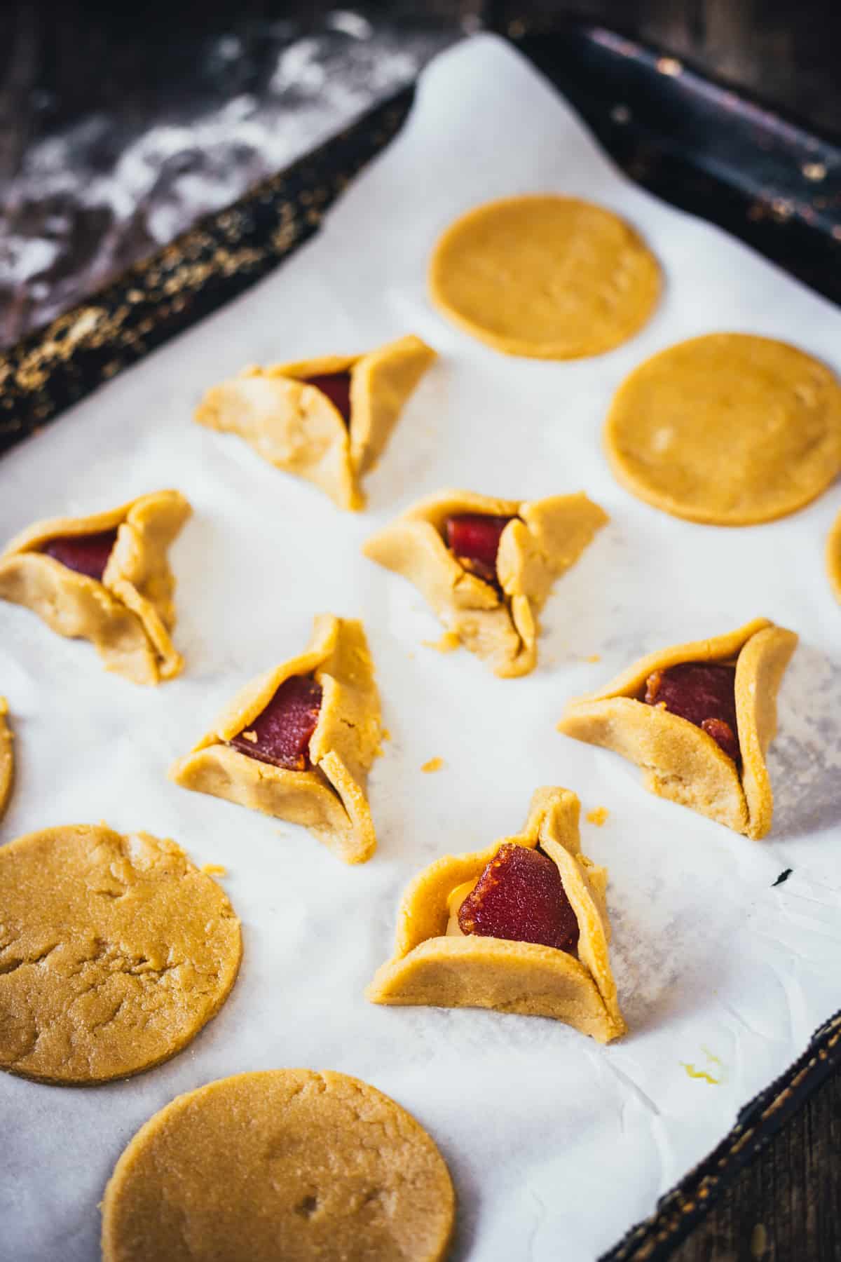 Unbaked guava and cheese hamantaschen cookies arranged on a parchment paper, with some cookies in a triangular shape and one appearing as a circle of dough.