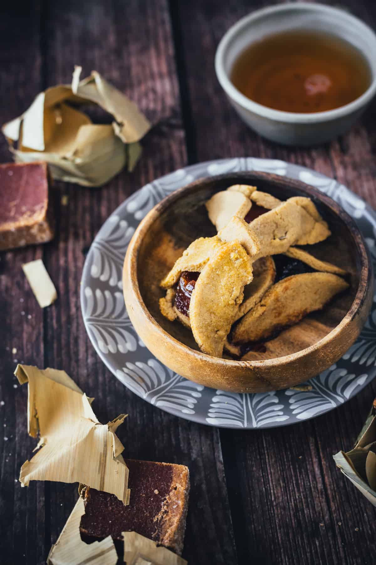 A bowl of bocadillo hamantaschen on a patterned plate, accompanied by a cup of tea and Hamantaschen Recipe sweets on a rustic wooden table.