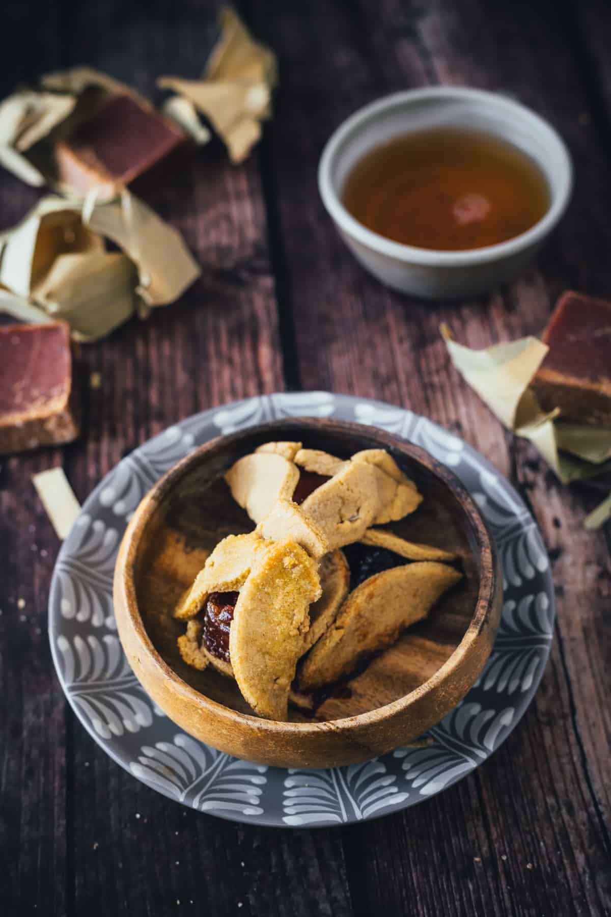 A bowl of freshly baked cookies and Cheese Hamantaschen served on a patterned plate with pieces of chocolate and a cup of tea in the background, displayed on a rustic wooden surface.