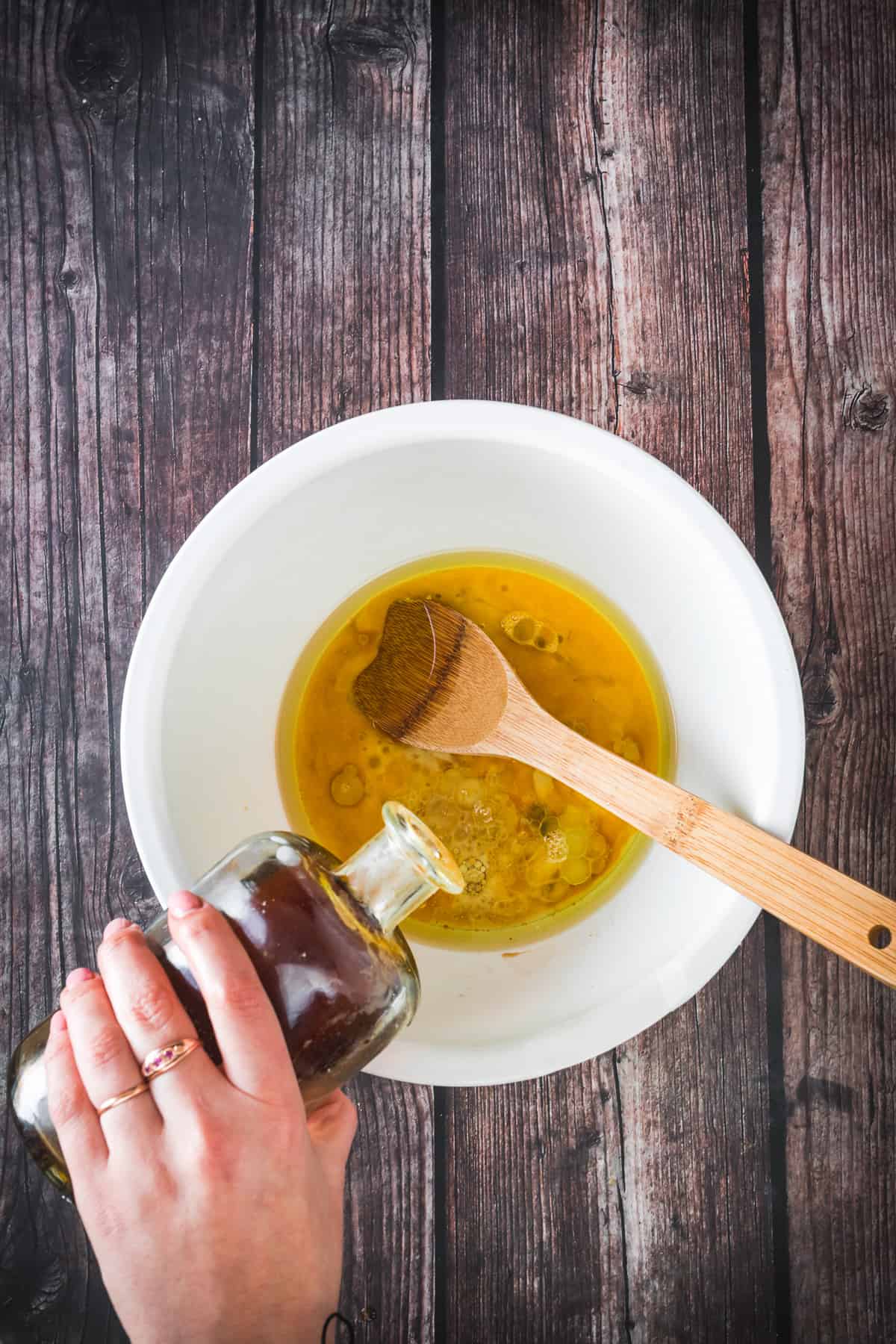A person pouring oil into a bowl with a honey and Bocadillo Guava mixture, using a wooden spoon for stirring, on a rustic wooden surface.