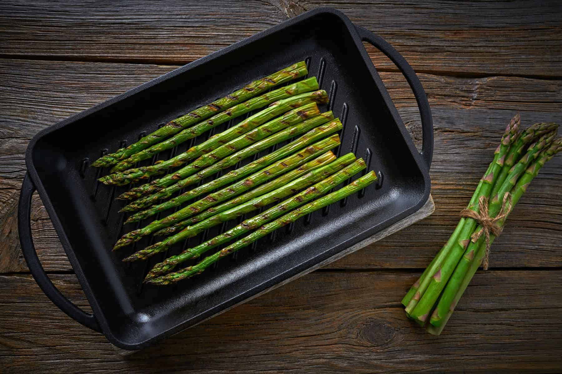 Fresh asparagus spears arranged neatly on a black roasting pan, ready to cook, with a bundle of asparagus placed beside it on a rustic wooden surface.