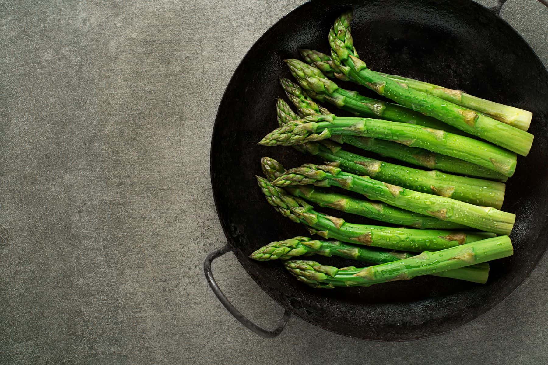 Fresh asparagus spears ready to cook in a cast iron skillet on a textured gray surface, perfect for exploring asparagus recipes.