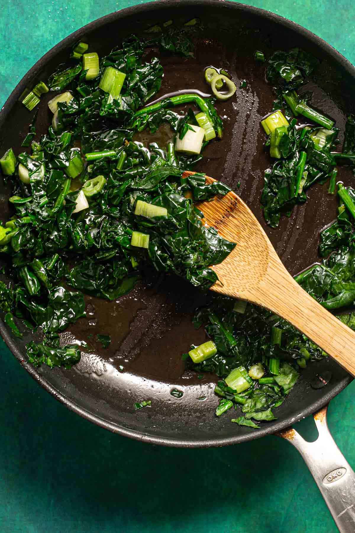 sauteed Spinach and green onions in a pan with a wooden spoon.