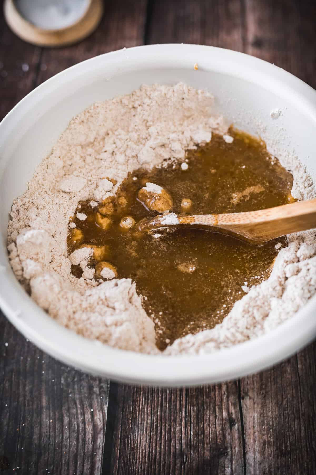 A wooden spoon is mixing wet ingredients into dry ingredients in a white bowl during a baking preparation process for Pistachio Hamantaschen.