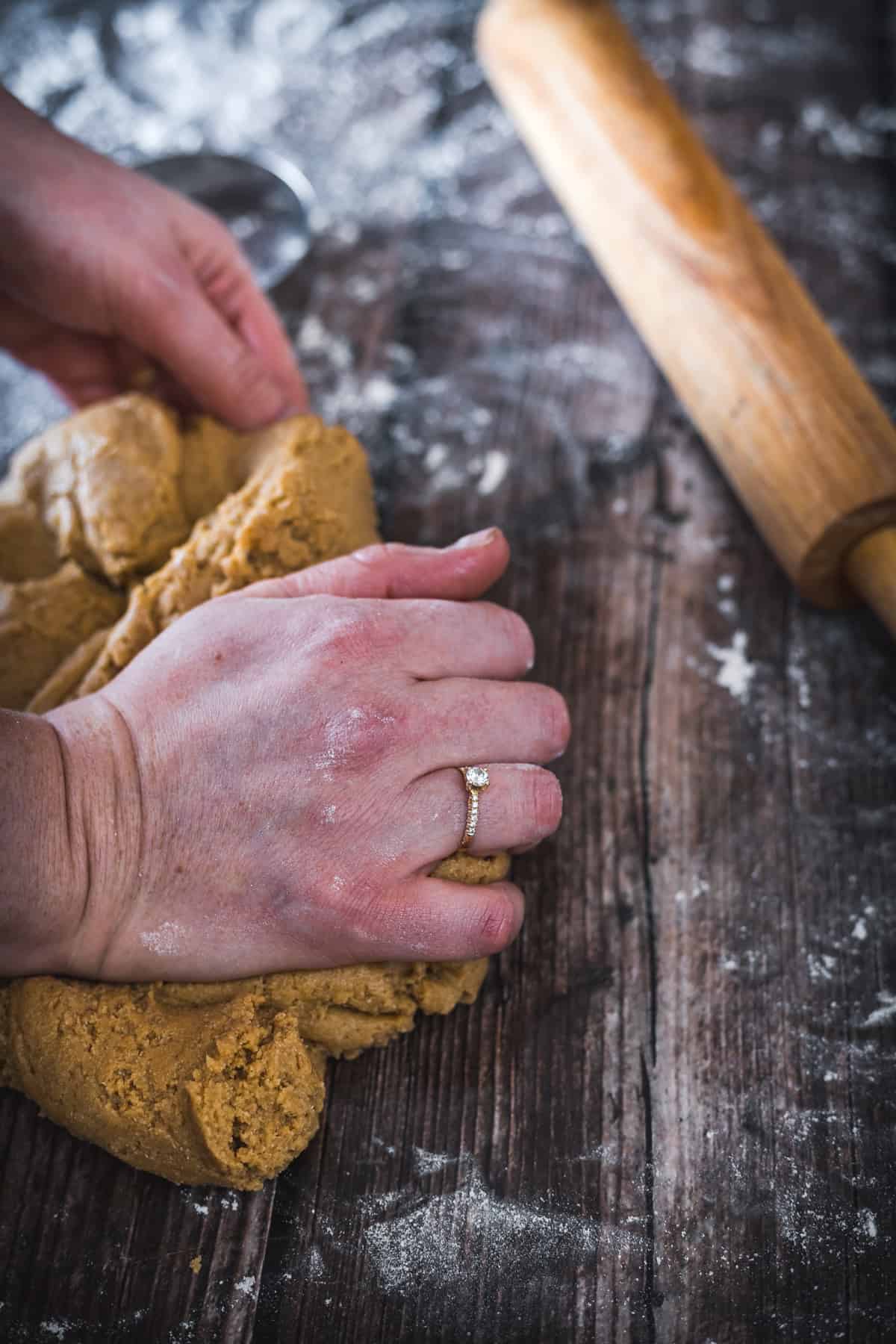 A person kneading dough on a wooden surface dusted with flour, with a rolling pin resting nearby, preparing to shape hamantaschen.