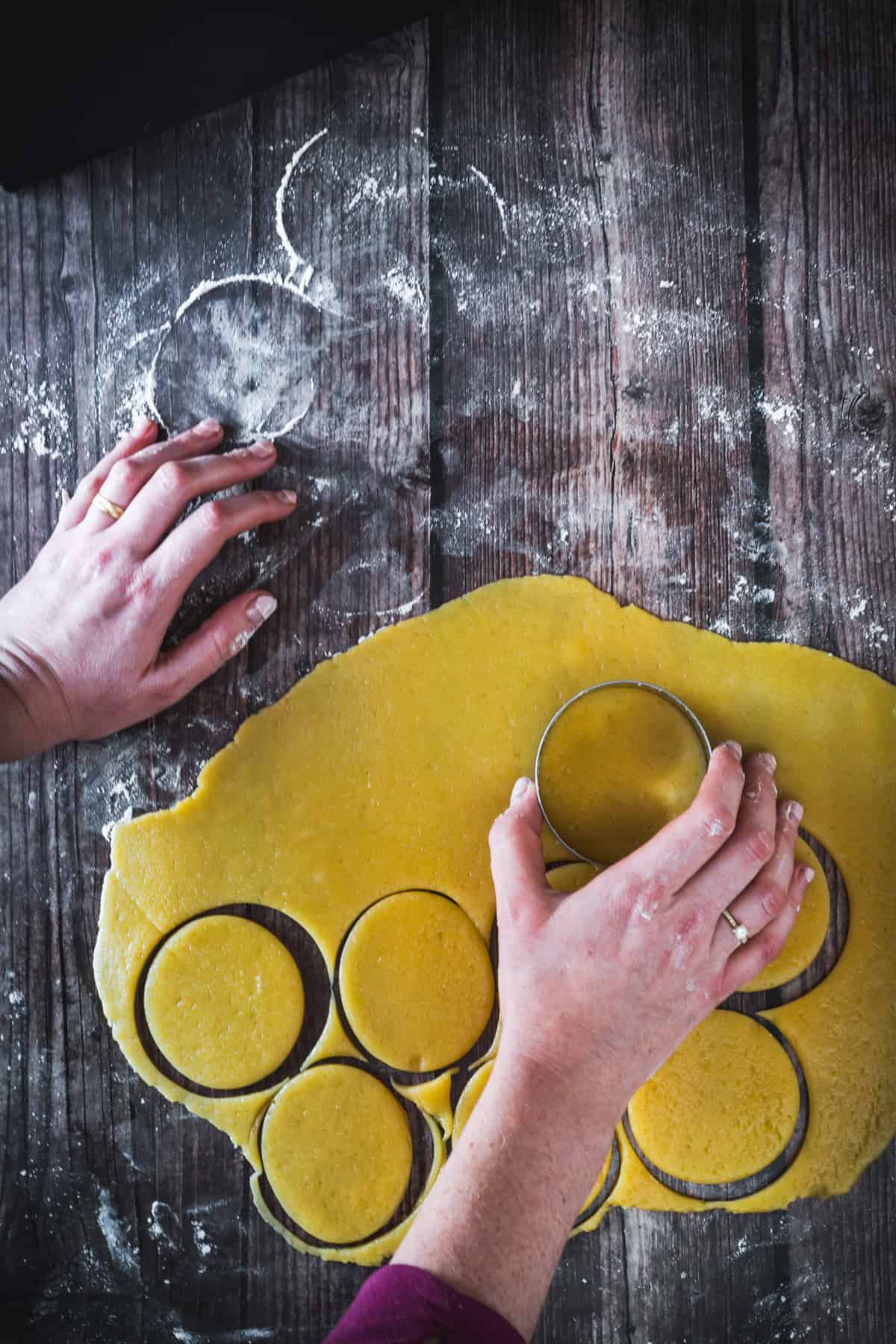 Person using a round cutter to shape rose pistachio Hamantaschen dough on a floured wooden surface.