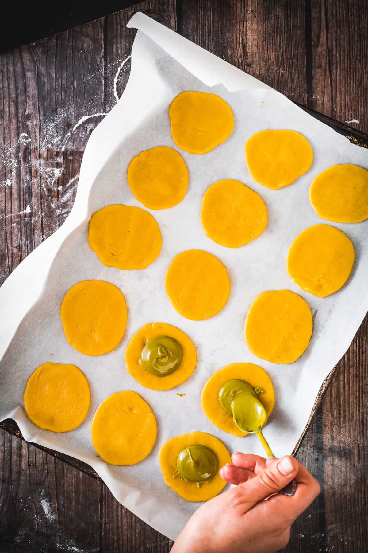 A person pipetting pistachio liquid onto circular orange dough laid out on baking paper.