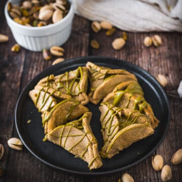 A black plate containing slices of Rose Pistachio-flavored cake with drizzled icing, surrounded by scattered pistachio nuts, on a wooden table.