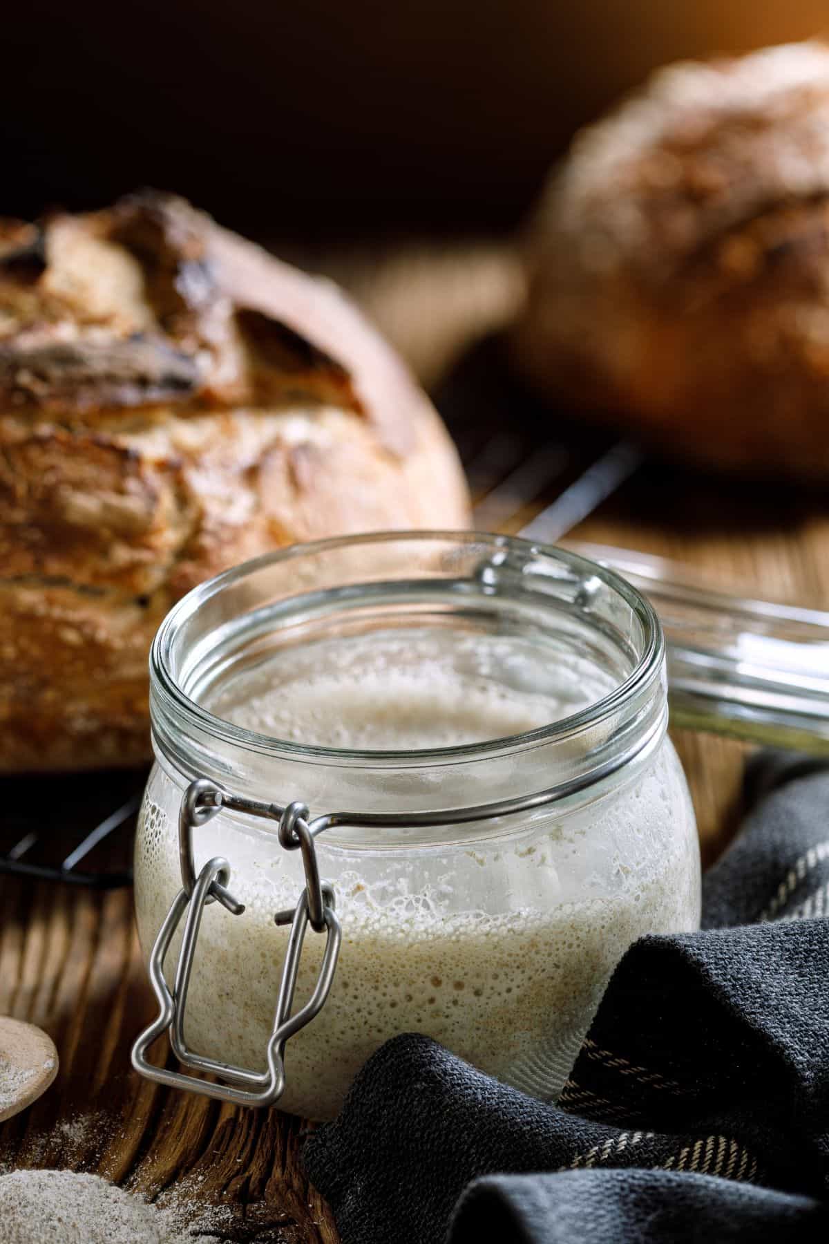 Glass jar containing traditional sourdough starter, with a loaf of freshly baked bread in the background.