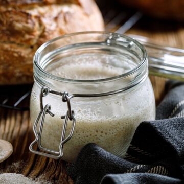 A glass jar filled with active traditional sourdough starter, situated on a wooden surface with a cloth napkin and freshly baked bread loaf in the background.